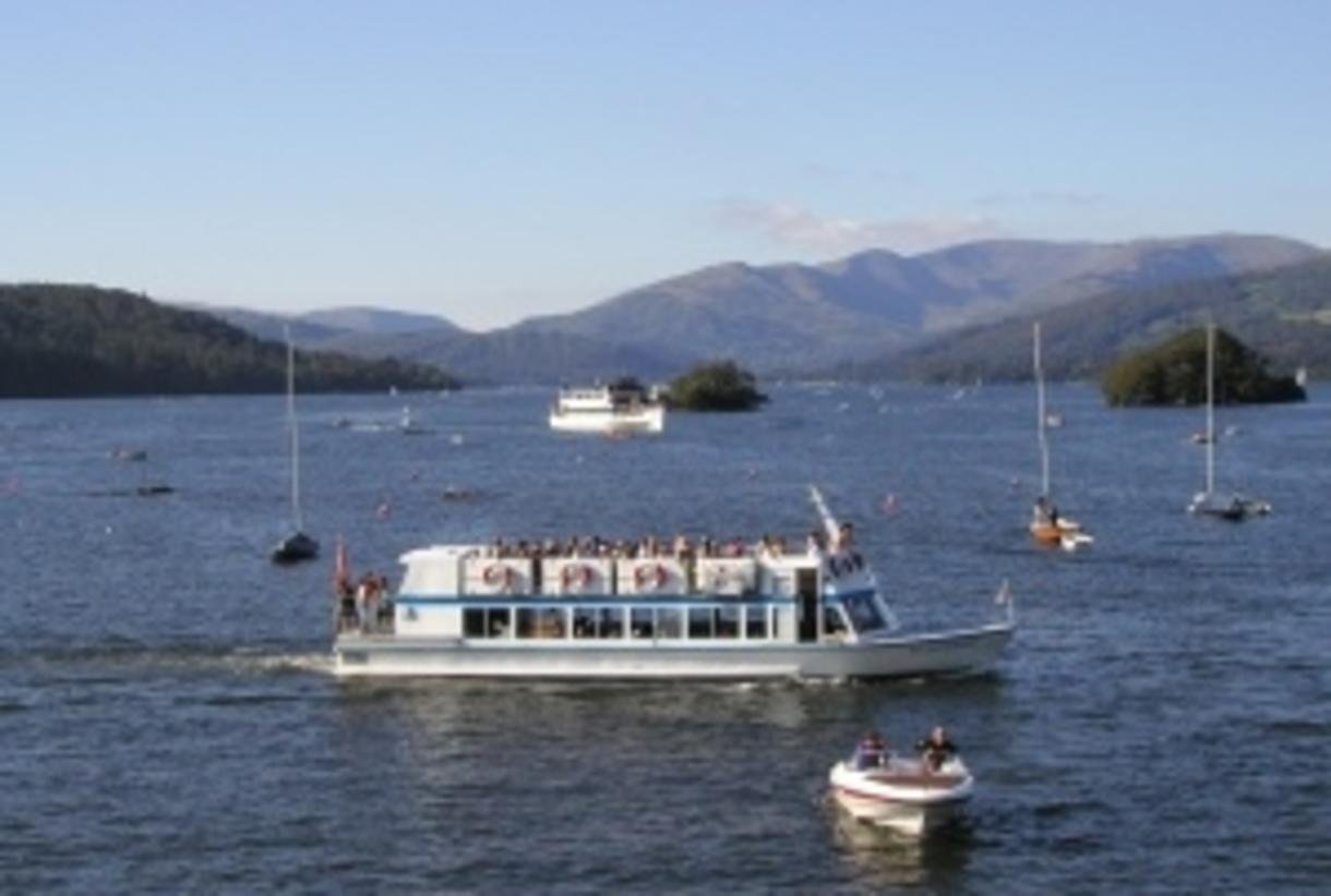 White two-decker tour boat with passengers cruising on a calm lake, with hills in the background and several small sailboats nearby. - Manchester Airport Taxi Transfer - Manchester Airport Taxi Transfer