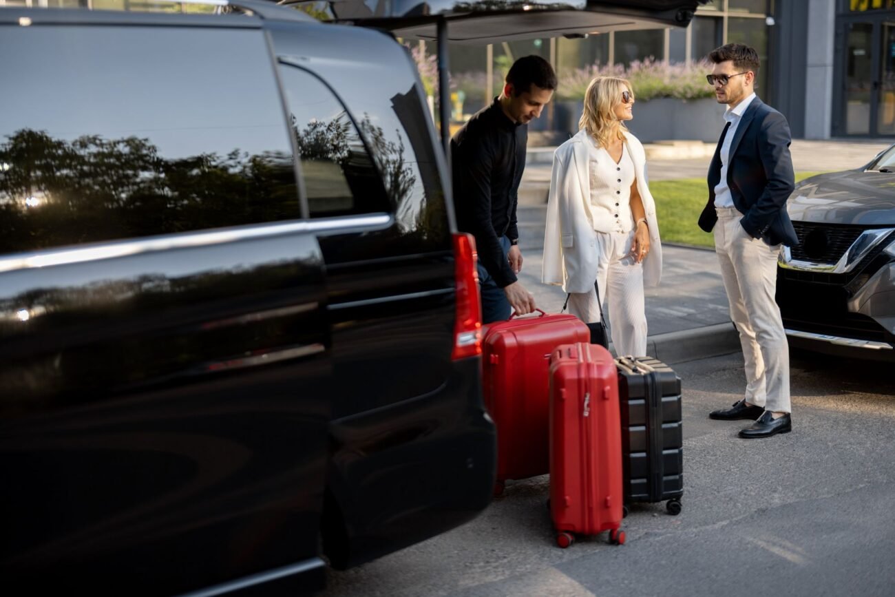 Three professionals with red and black luggage stand near a black van outside a modern building, ready to travel. - Manchester Airport Taxi Transfer - Manchester Airport Taxi Transfer