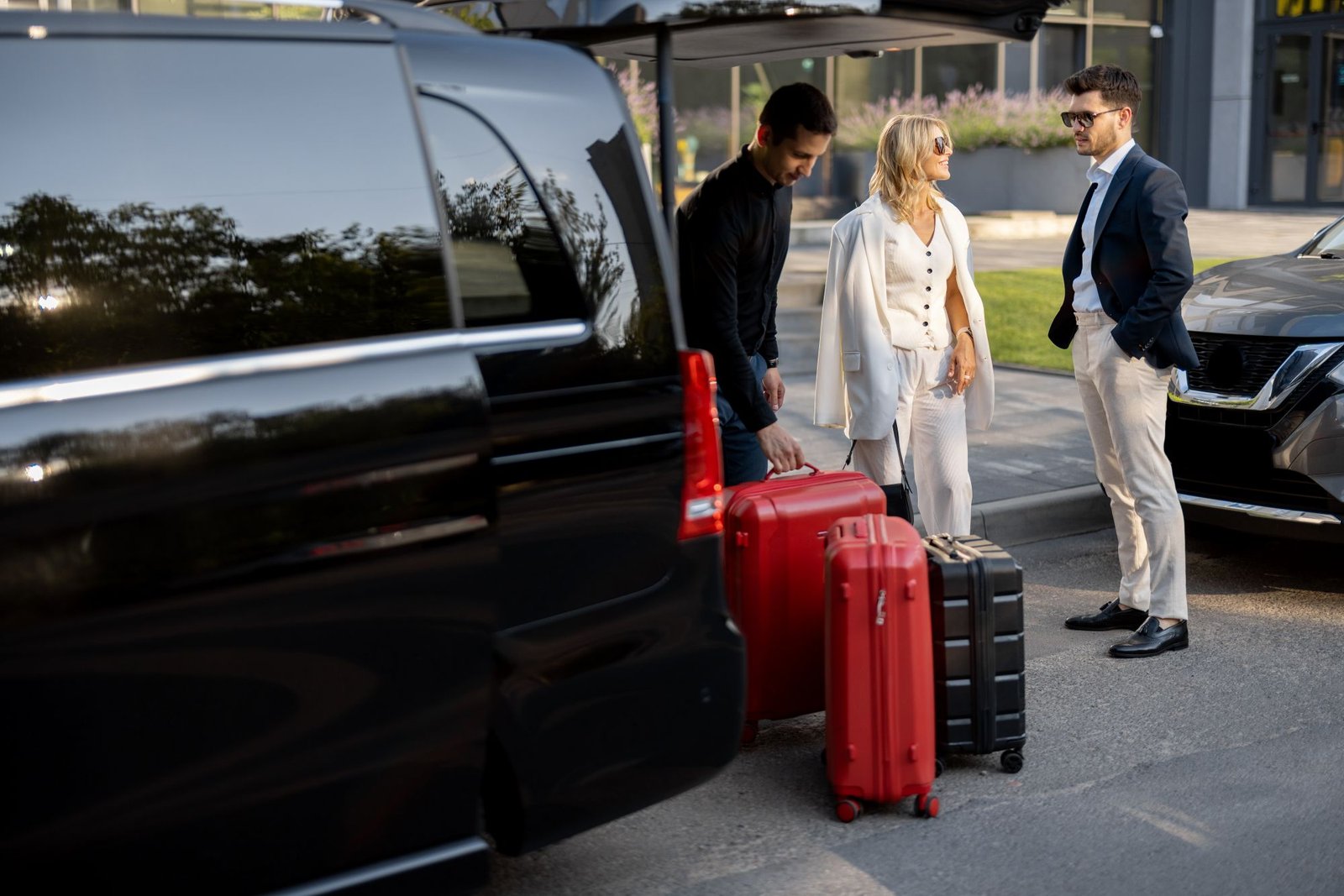 Three professionals with red and black luggage stand near a black van outside a modern building, ready to travel. - Manchester Airport Taxi Transfer - Manchester Airport Taxi Transfer
