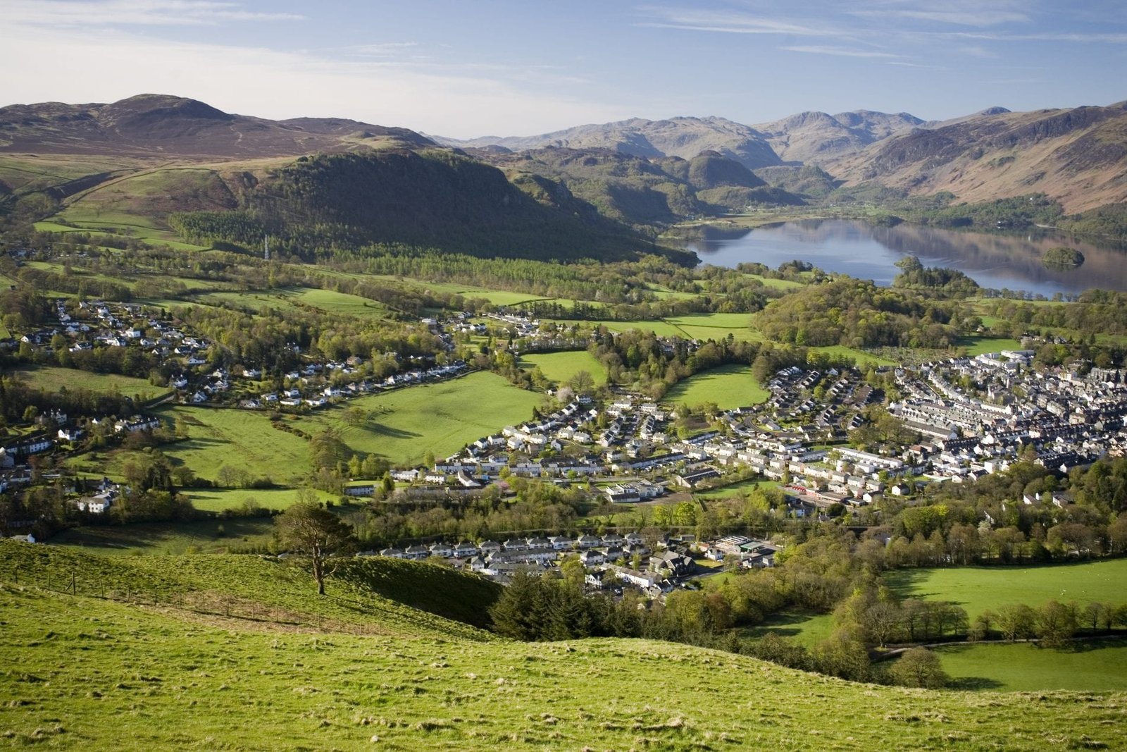 A valley town sprawled across green fields with a lake and distant mountains under a clear sky. - Manchester Airport Taxi Transfer - Manchester Airport Taxi Transfer