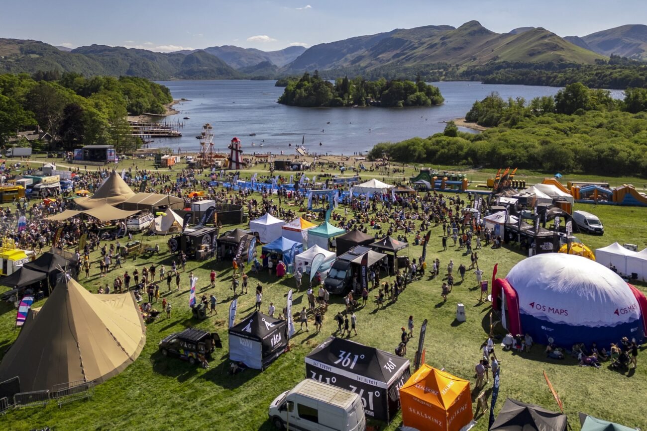 Aerial view of a large lakeside festival with numerous tents, stalls, and attendees on a grassy field by a calm lake and green hills in the distance. - Manchester Airport Taxi Transfer - Manchester Airport Taxi Transfer