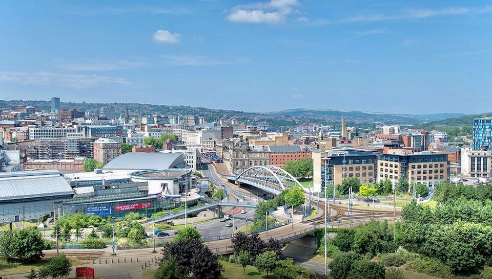 Bird's-eye view of a city with a river, arched pedestrian bridge, and modern buildings in the distance. - Manchester Airport Taxi Transfer - Manchester Airport Taxi Transfer