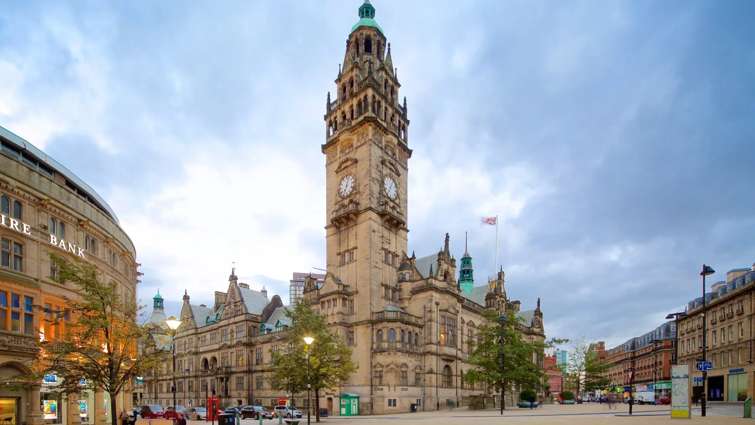 Historic city square with a tall Gothic clock tower rising above ornate stone buildings and a cloudy sky, street activity below. - Manchester Airport Taxi Transfer - Manchester Airport Taxi Transfer