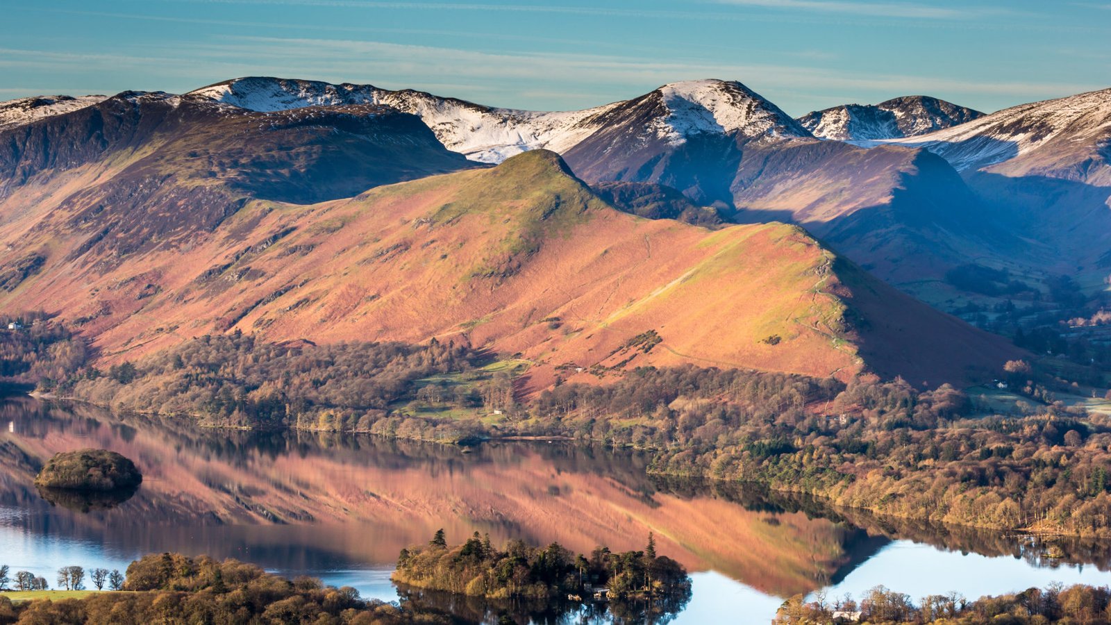 Snow-dusted mountain range with a calm lake reflecting the pinkish-brown slopes and blue sky in a tranquil valley. - Manchester Airport Taxi Transfer - Manchester Airport Taxi Transfer