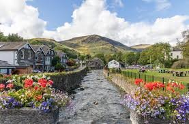 Colorful flower-filled canal in a village, with stone walls, row houses, and green hills in the background. - Manchester Airport Taxi Transfer - Manchester Airport Taxi Transfer