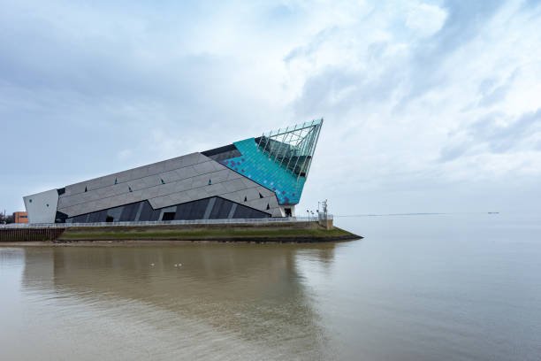 Modern angular building jutting into calm water, with teal glass roof and grey slanted walls on a narrow shoreline. - Manchester Airport Taxi Transfer - Manchester Airport Taxi Transfer