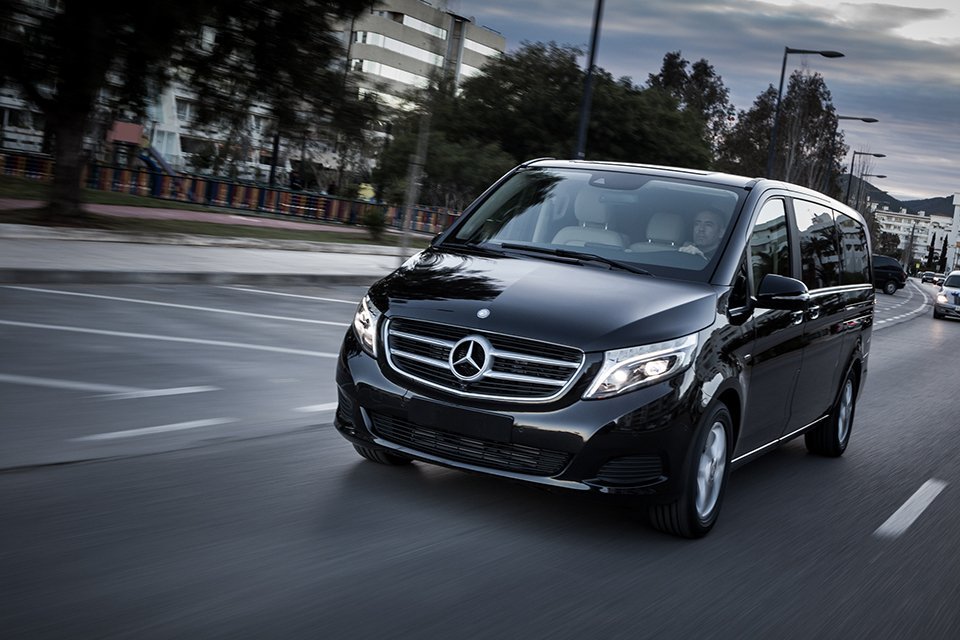Black Mercedes-Benz van driving on a city street, front-right angle, headlights on. - Manchester Airport Taxi Transfer - Manchester Airport Taxi Transfer
